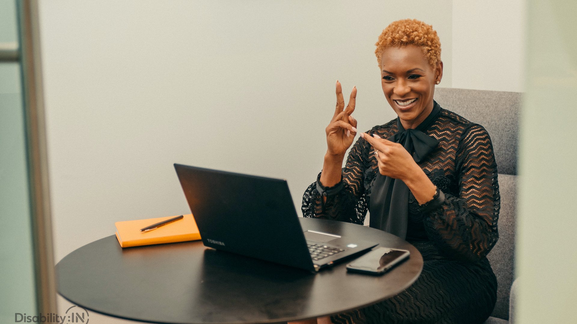 A woman communicates with sign language. She is sitting at a table with a laptop open in front of her.