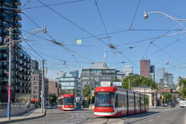 Photo of two modern streetcars coming to a corner. The current Toronto skyline, including an obstructed view of the CN tower, is in the background.