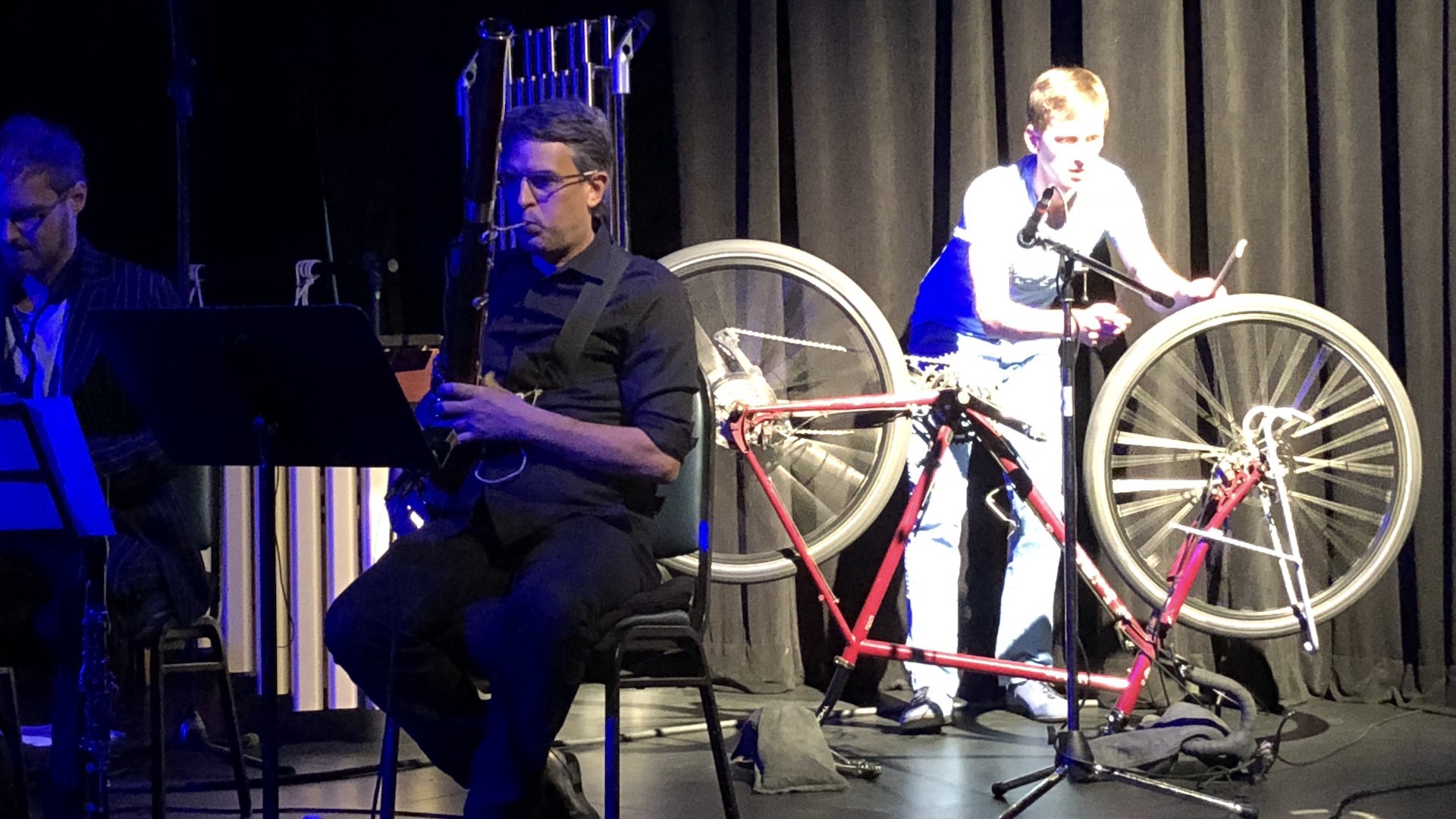 Man sits and plays a musical instrument. Another man stands behind him and makes music using a bicycle tire.