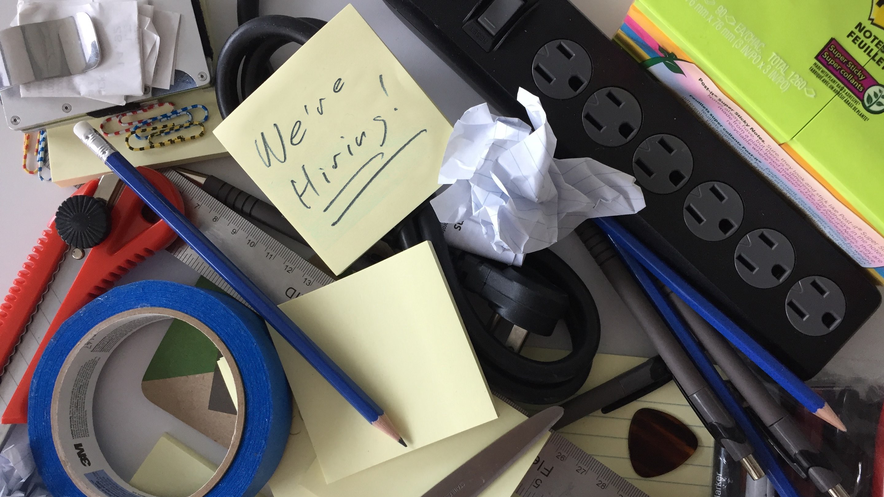 a pile of tape, a box cutter, a power bar, post-it notes, pencil and a note reading We're hiring!