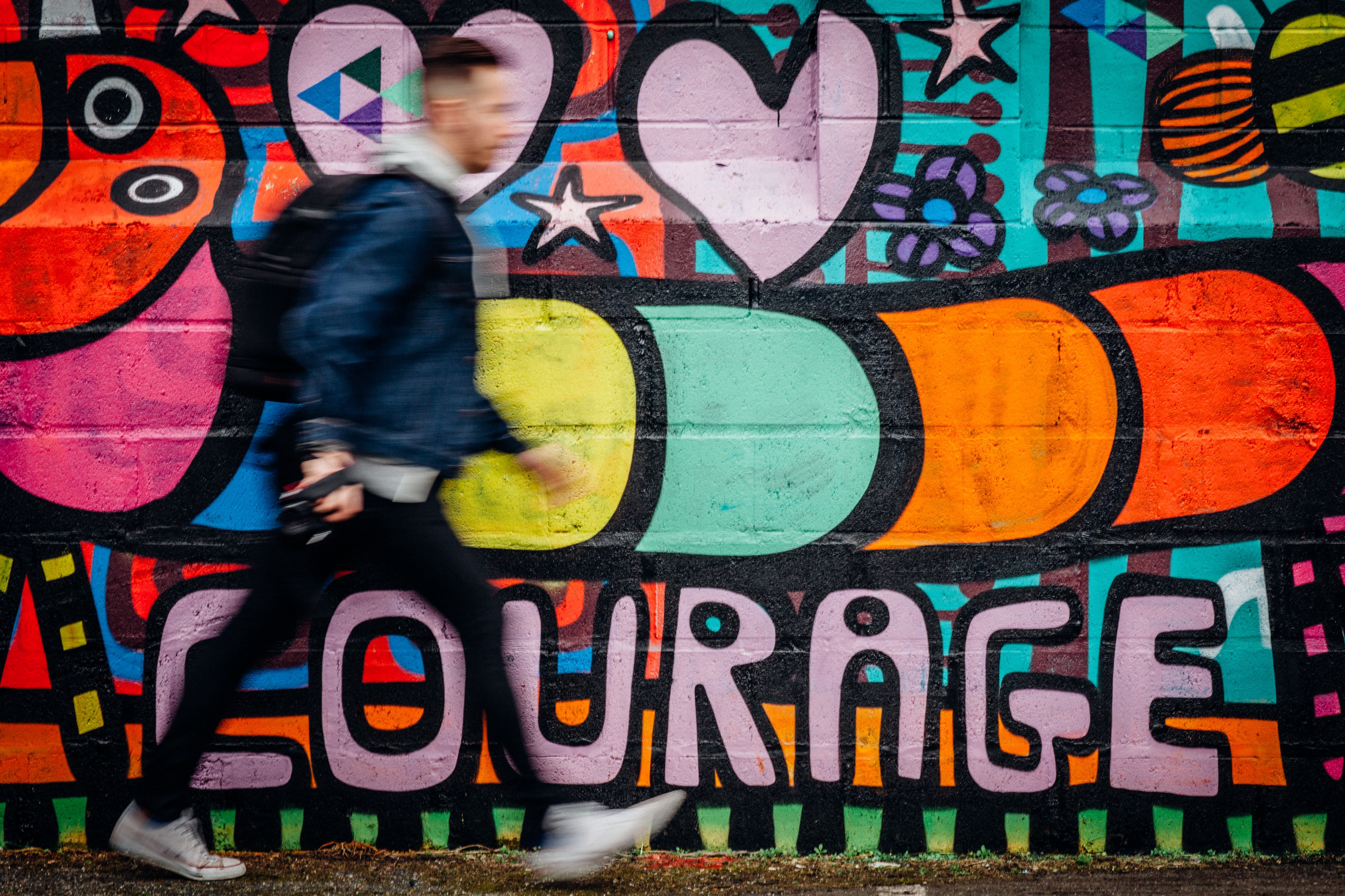 A blurred-person walking quickly in-front of a wall fo graffiti that reads 'Courage'