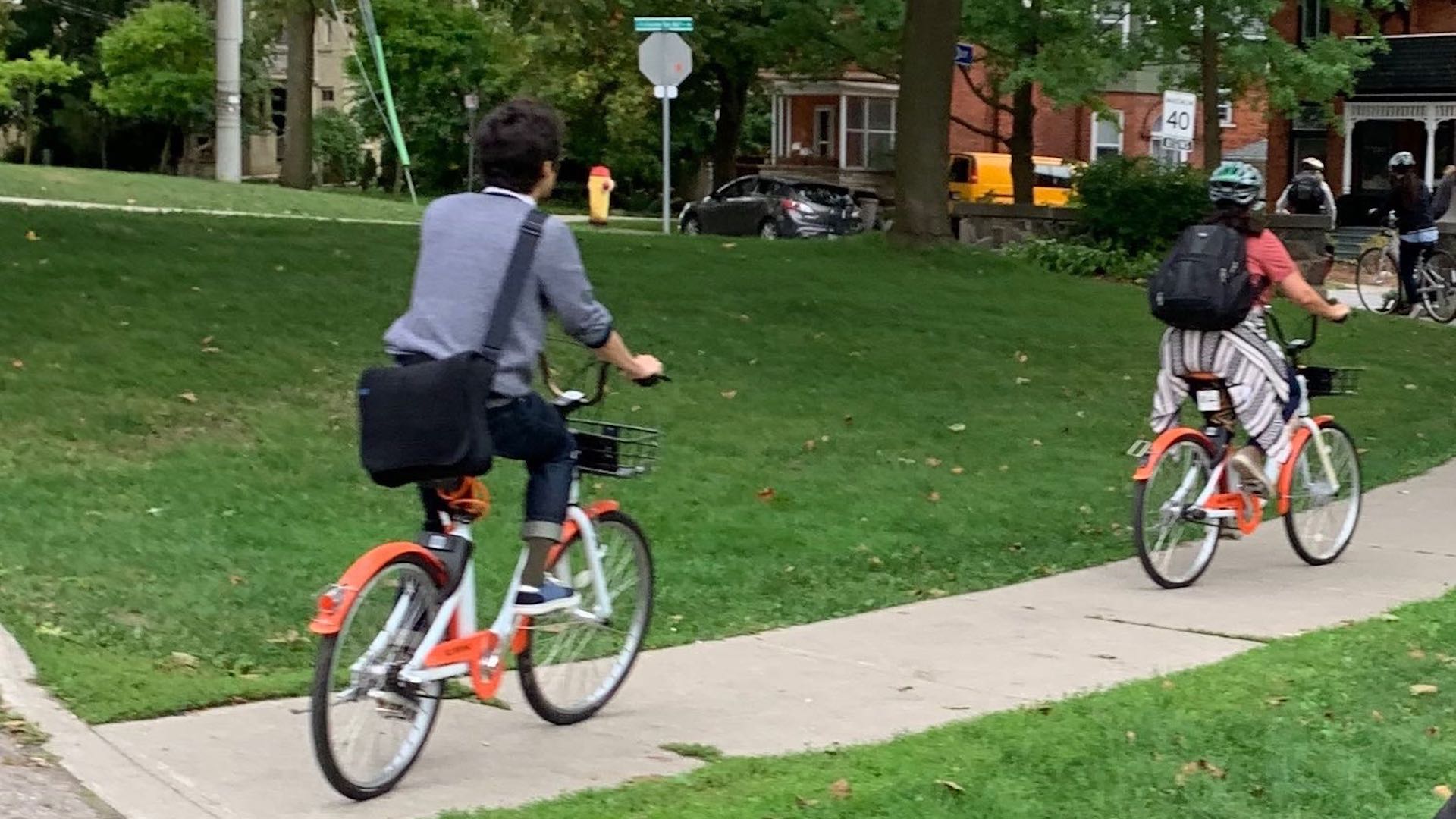 Two people ride Drop Mobility bikes along a sidewalk