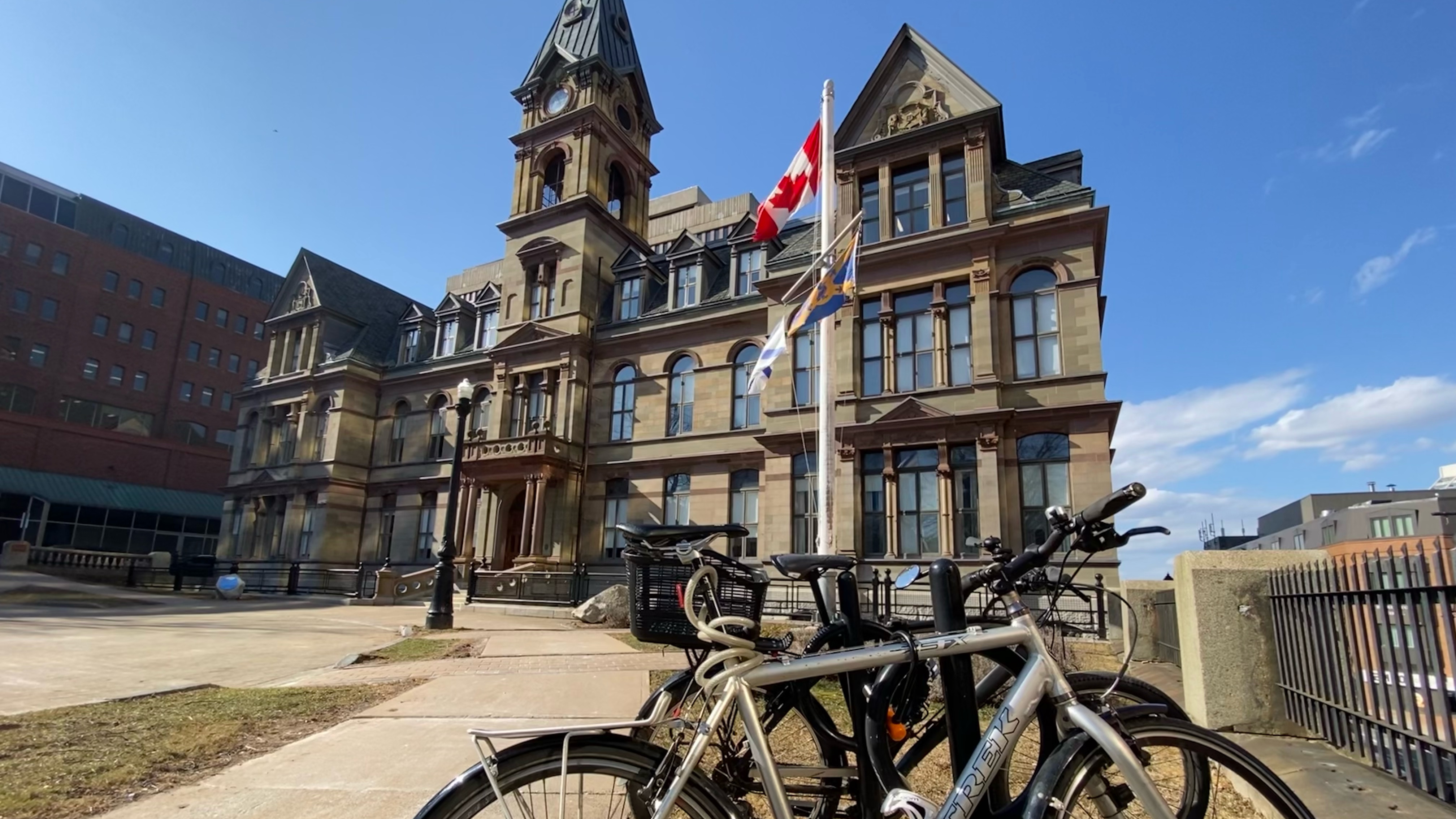 In the background there's an older stone building with a clock tower in the centre of it. In the middle ground there are flags on a flag pole. In the foreground there's a bike locked to a back rack. The sky is blue with just a few clouds.