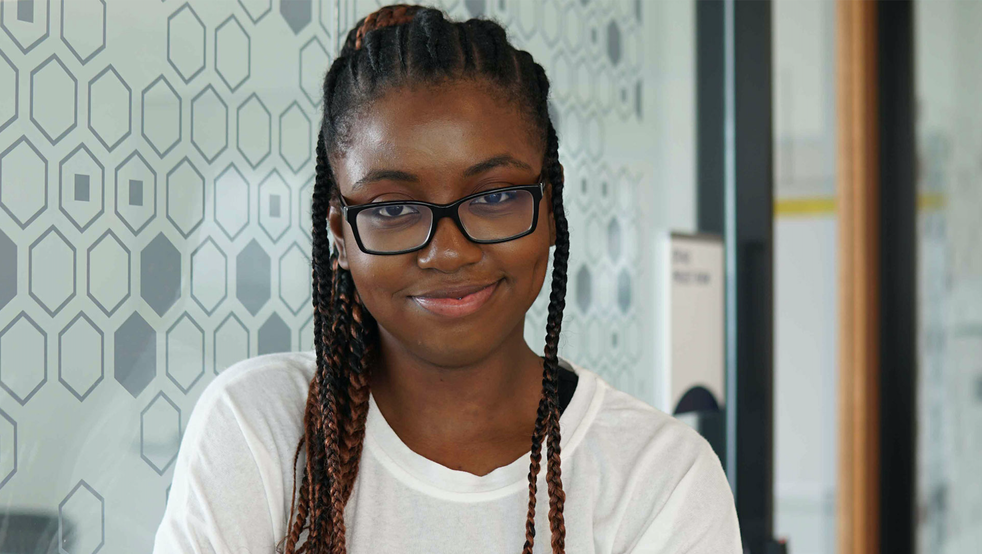 Kedice Jones is wearing glasses and a white shirt and is standing, arms crossed, in front of a wall with a hexagon pattern.