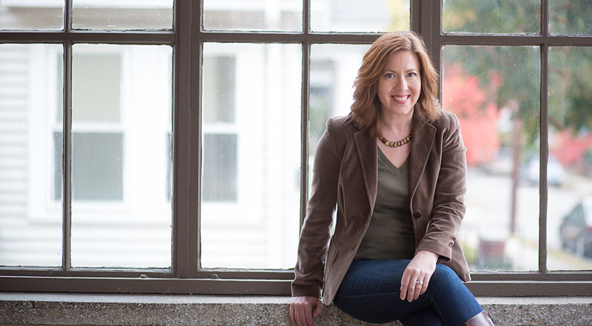 Content strategist Margot Bloomstein sits on a window ledge. She's wearing a brown blazer and jeans.