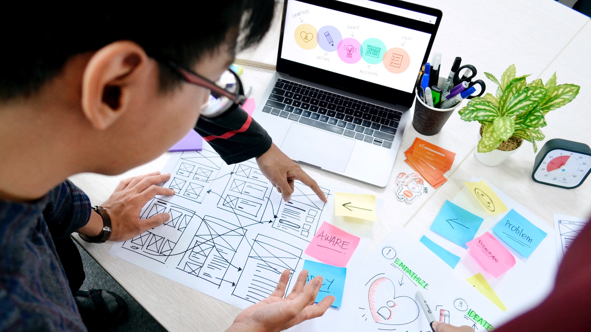 Hands pointing at paper prototypes on a desk. A laptop is open on the table and colourful sticky notes surround it.