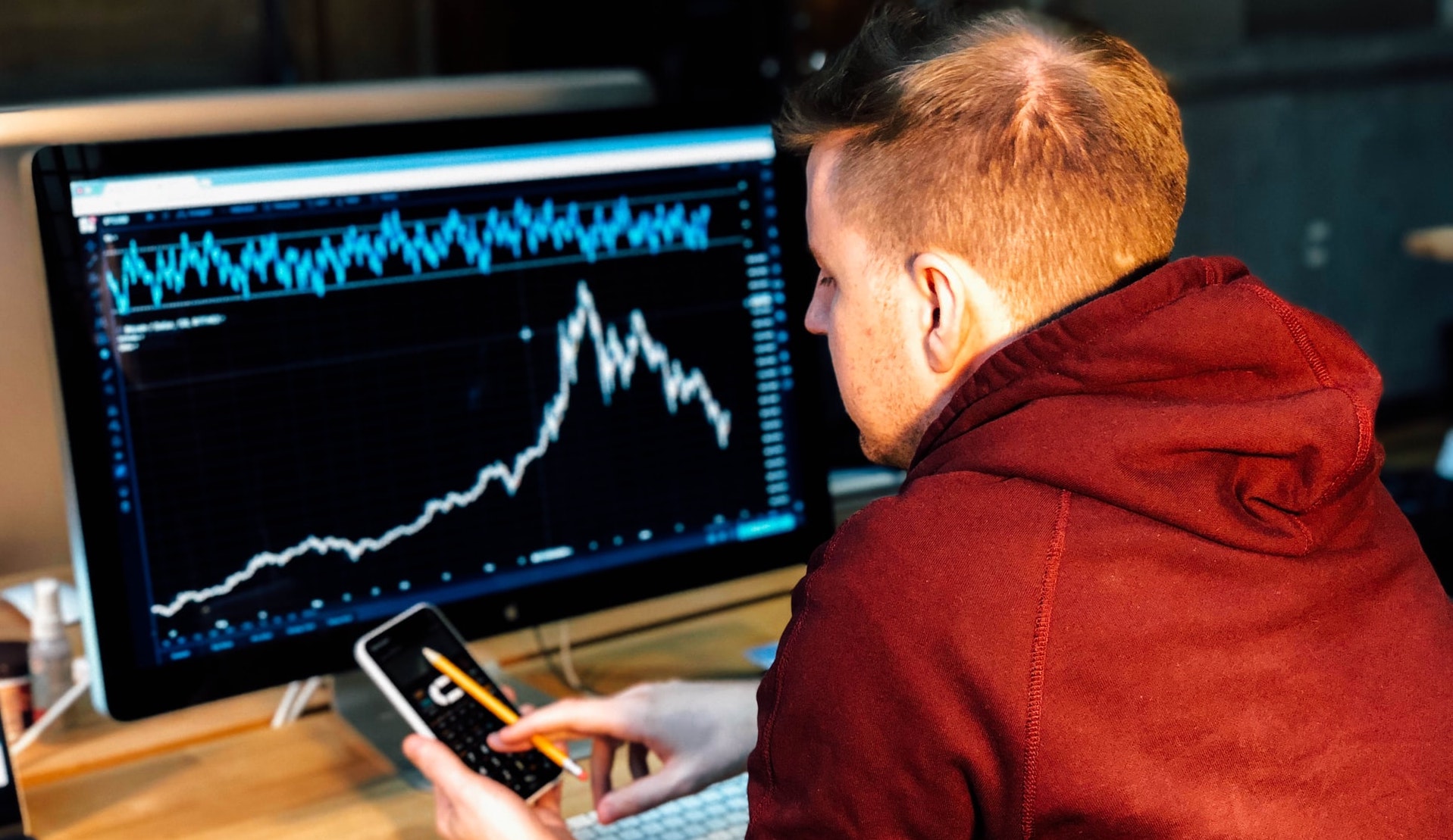 A man wearing a red hoodie and holding a cell phone sits in front of a computer screen showing a graph. 