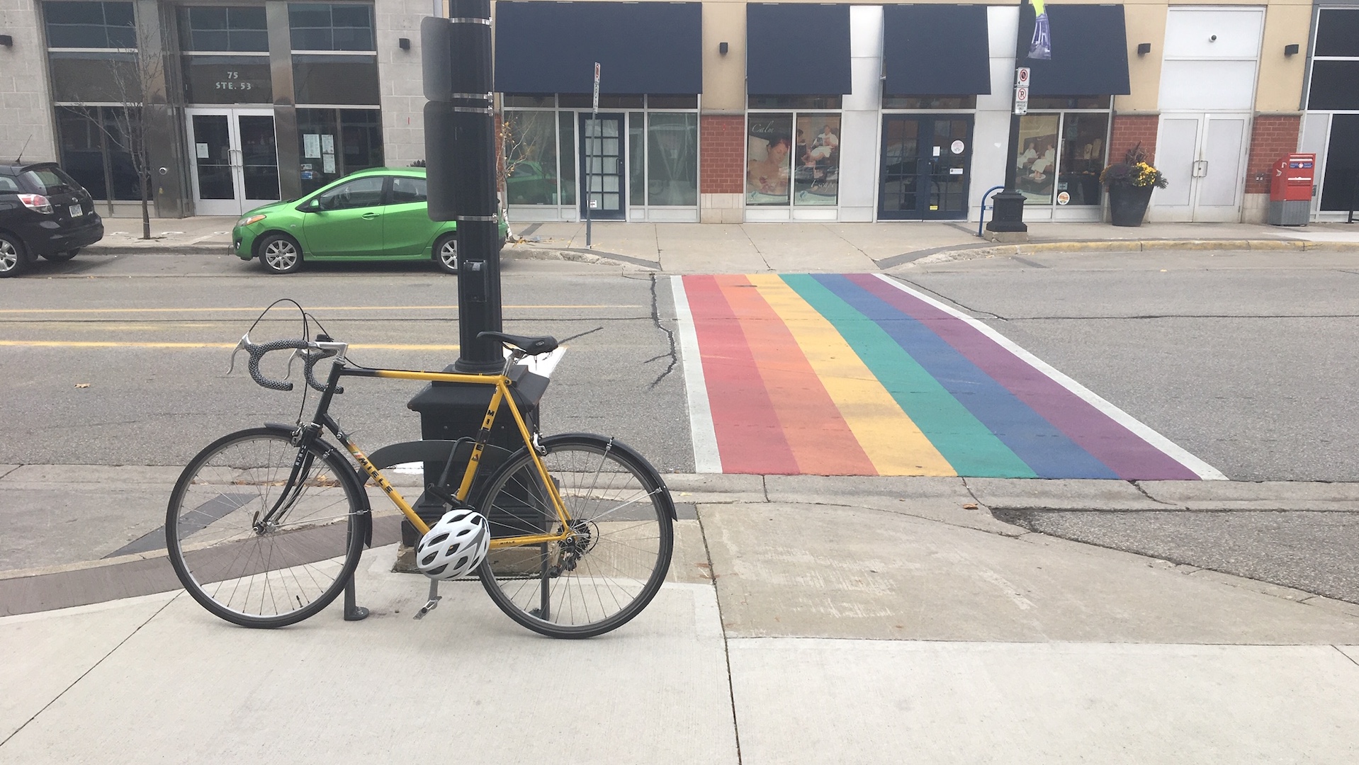 A crosswalk that has painted in rainbow colours. A bike is in the foreground locked to a bike rack with a helmet attached to the bike