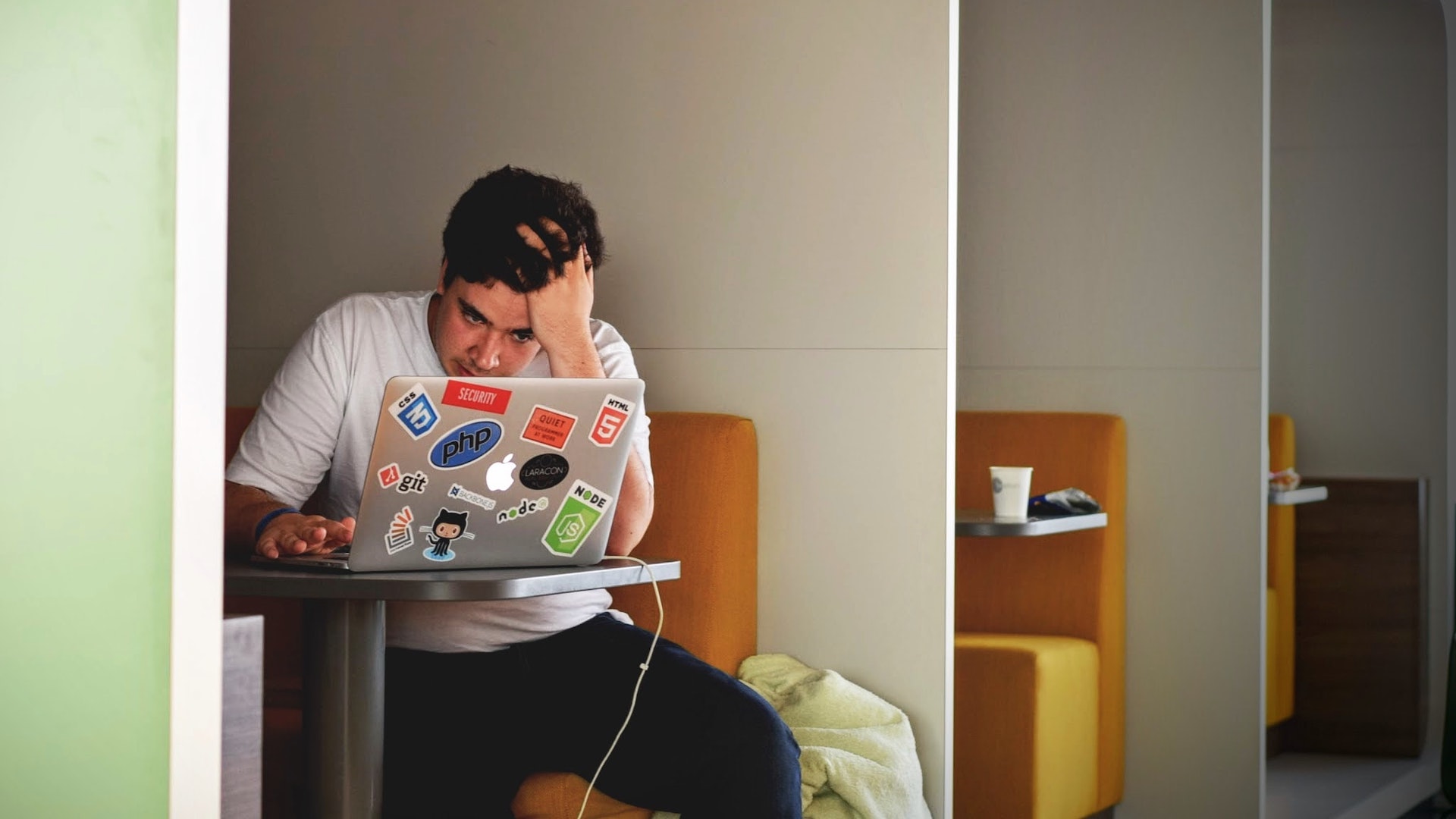 A stressed person sits in a booth at a tech company with a laptop open before him.