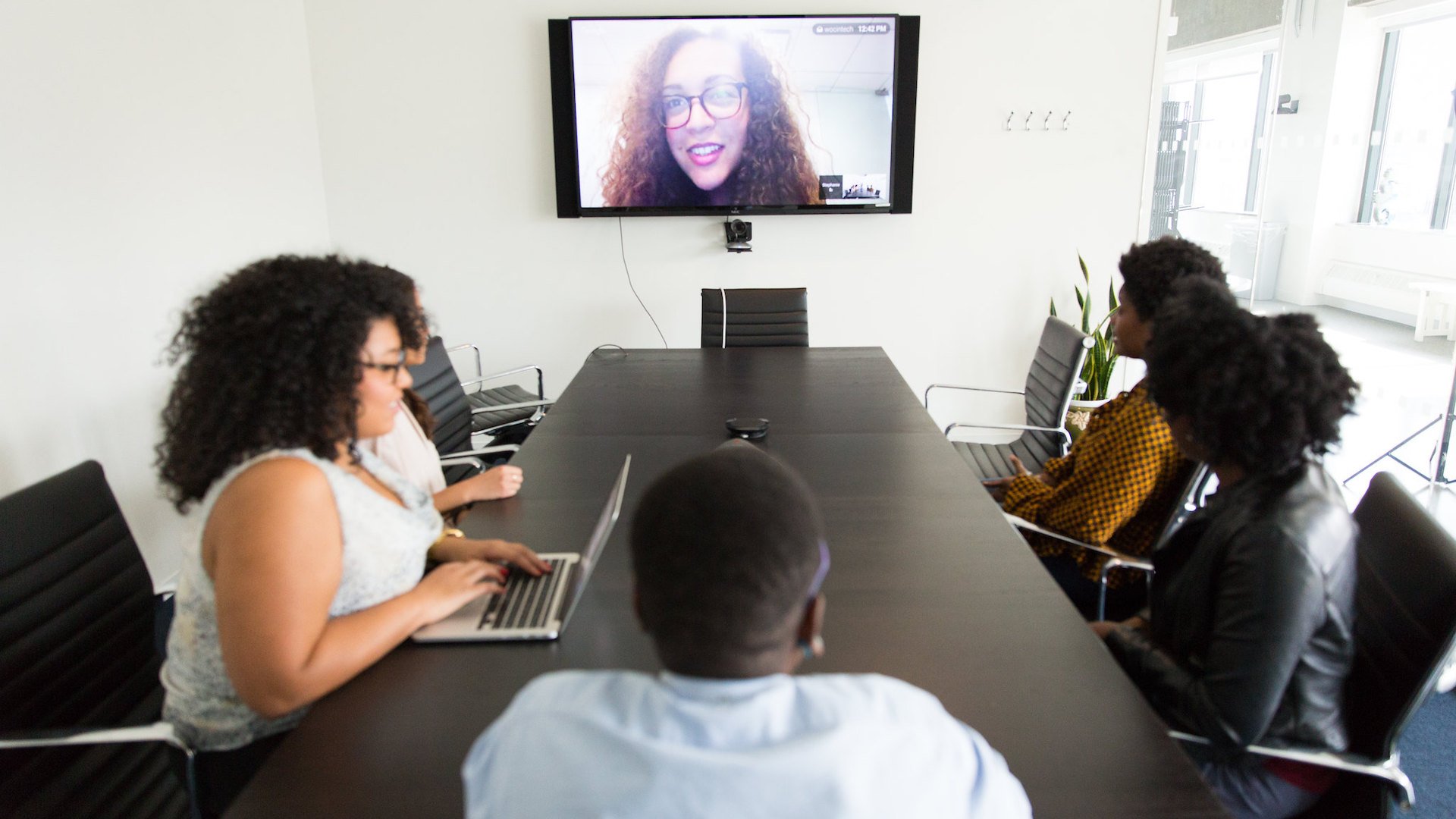 Five people sit around a boardroom table talking to a woman who appears via video.