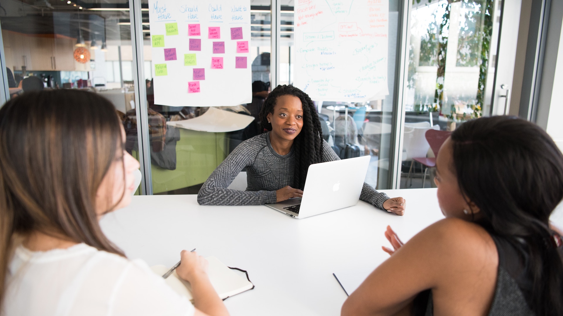 3 women in a discussion in a tech office, one has a laptop and there are post-its on a glass wall.