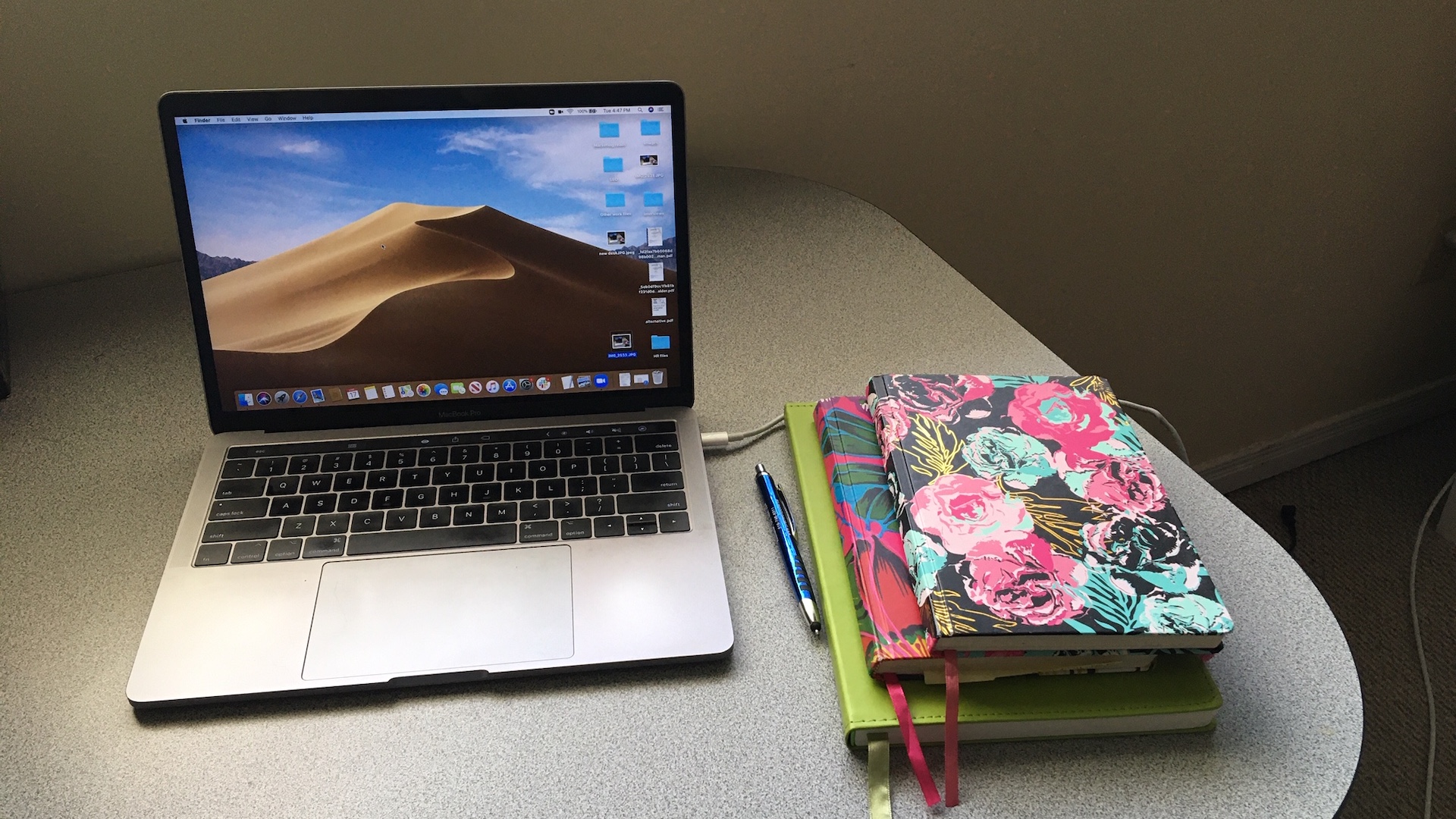 A MacBook sits open on a desk with several notebooks and a pencil.