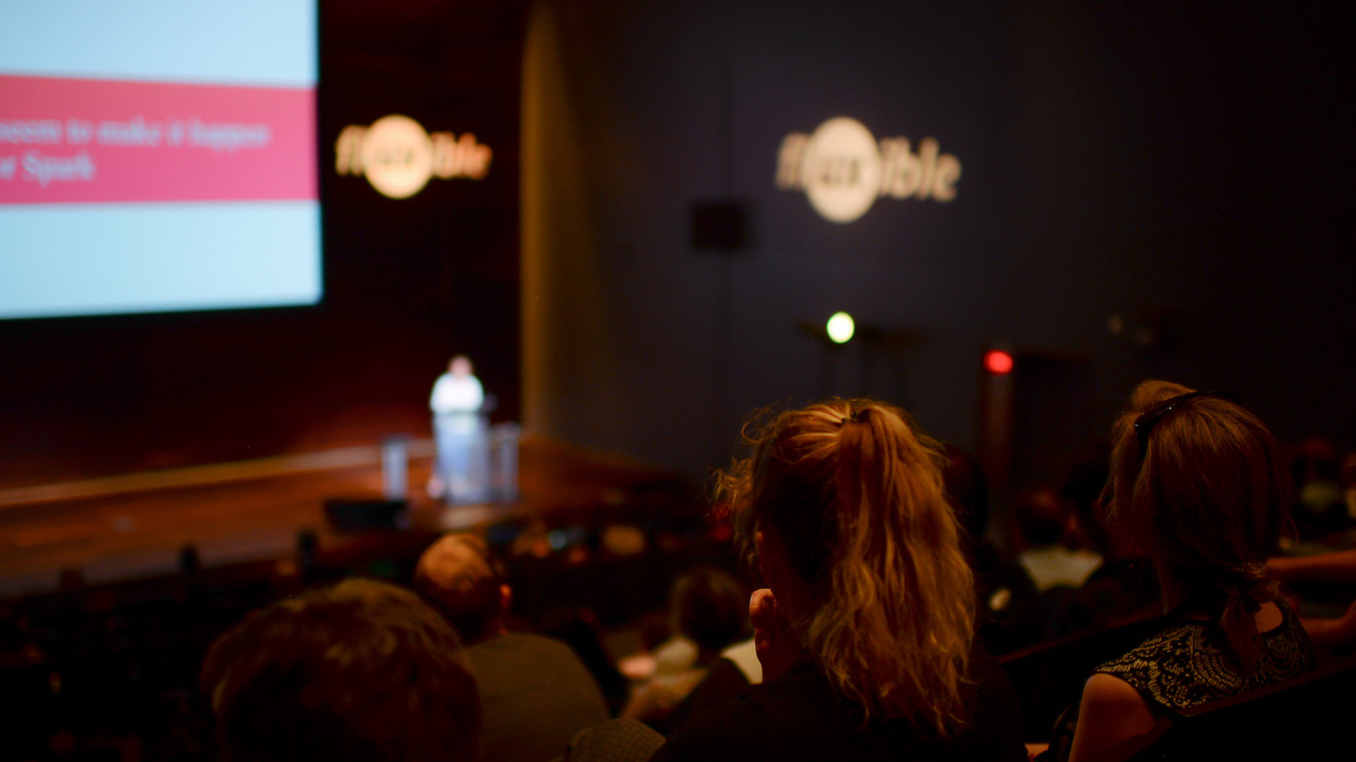 A person stands on the stage during Fluxible Conference while people from the audience look on.