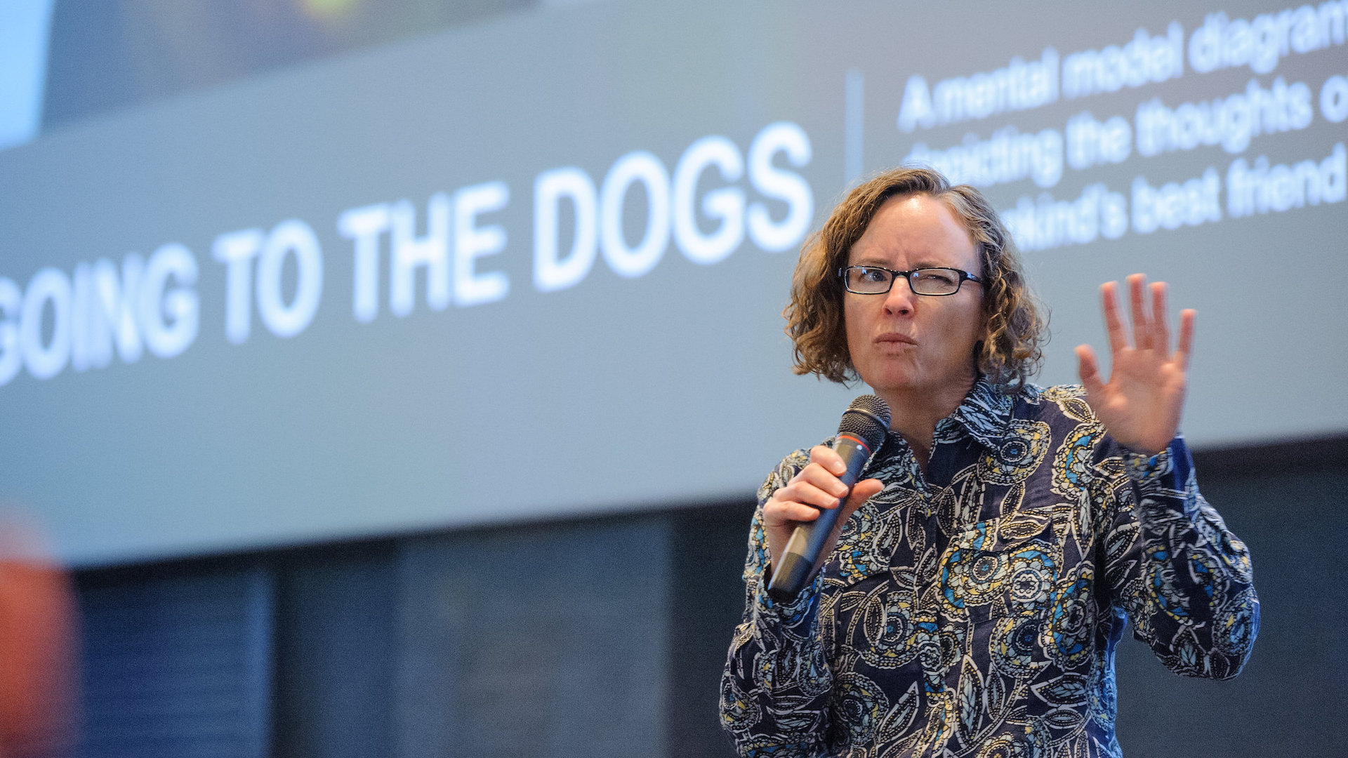 Indi Young squints her eye and speaks into a microphone while standing on a stage at Fluxible.