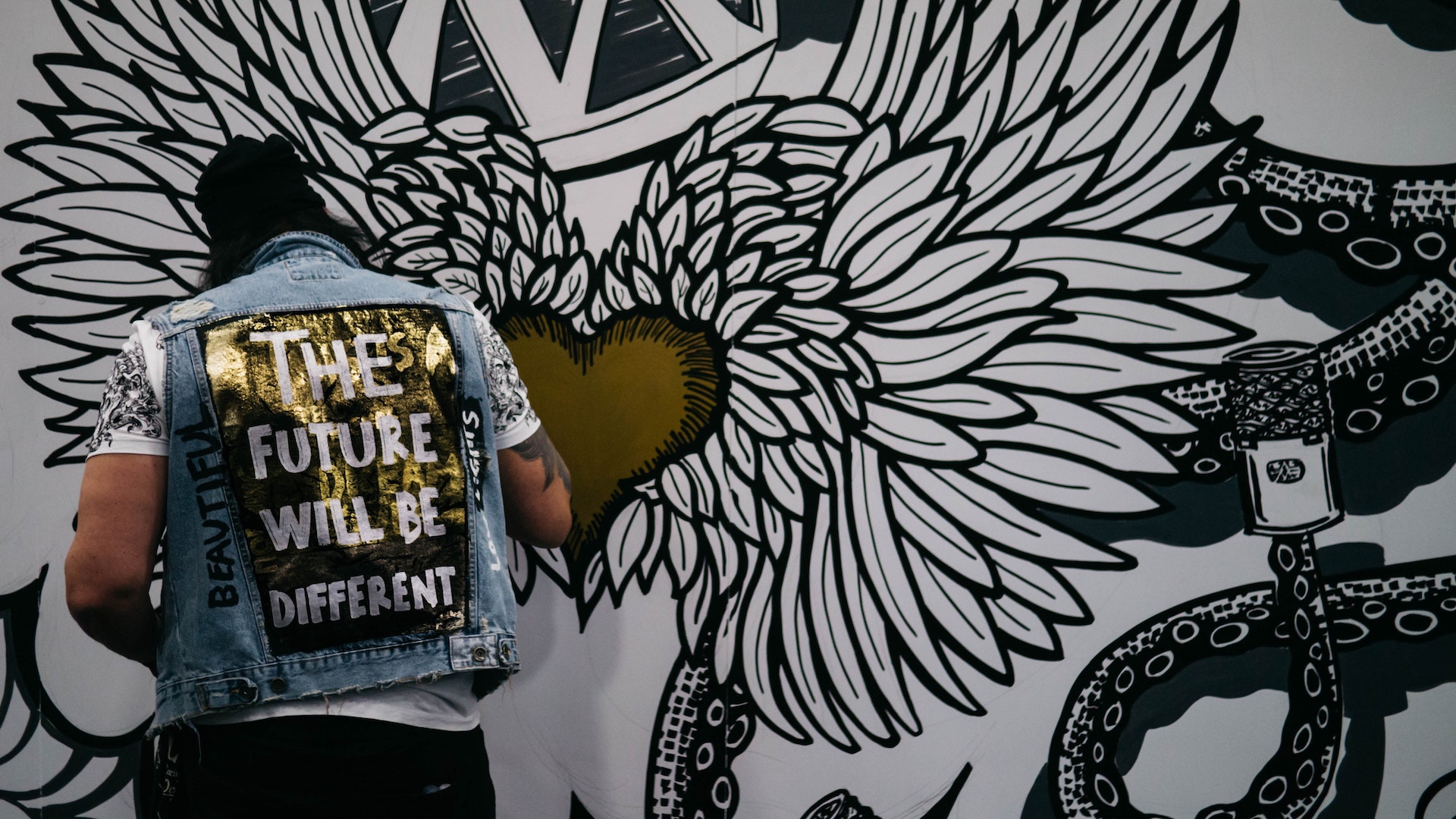 An artist paints a wall mural, his painted jean jacket reads 'the future will be different'.